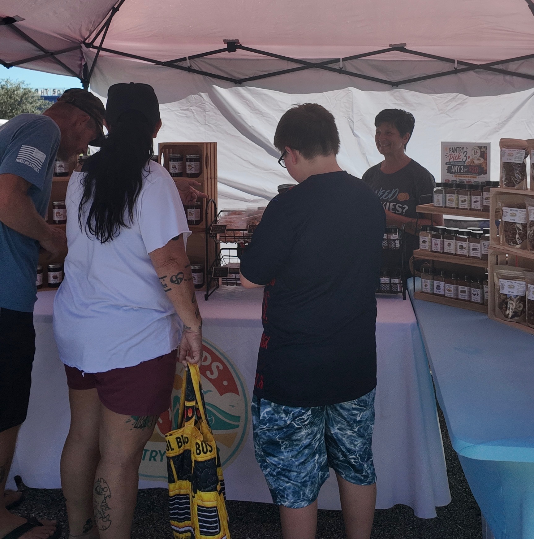 Shoppers browsing jars of jam, cookies and spice blends while a smiling vendor chats under a pink tent