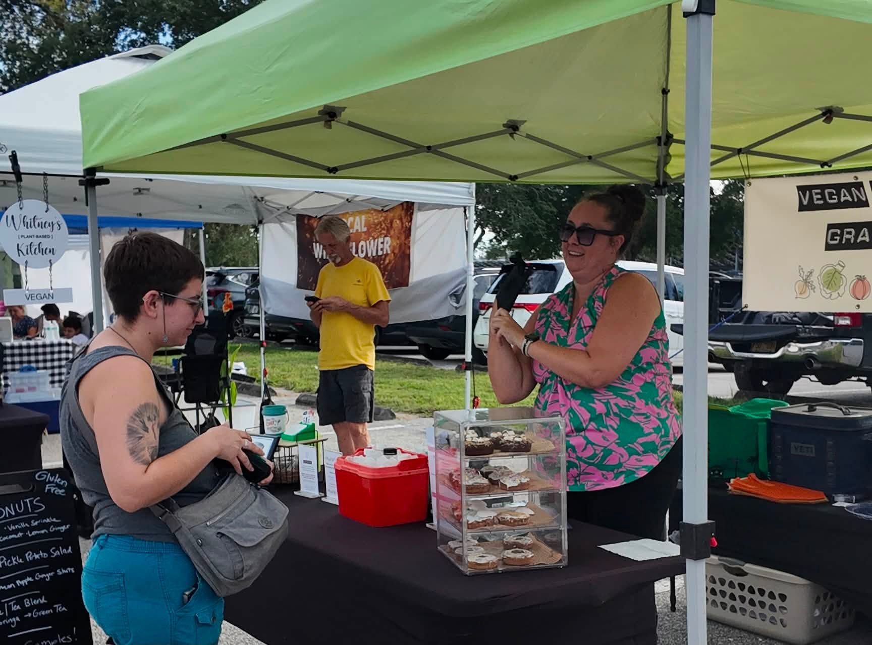 A shopper paying at a plant-based bakery tent with a case of frosted donuts