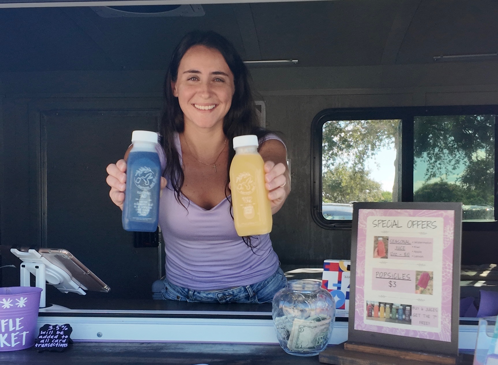 A smiling vendor holding out bottles of blue and yellow cold-pressed juice from her trailer window